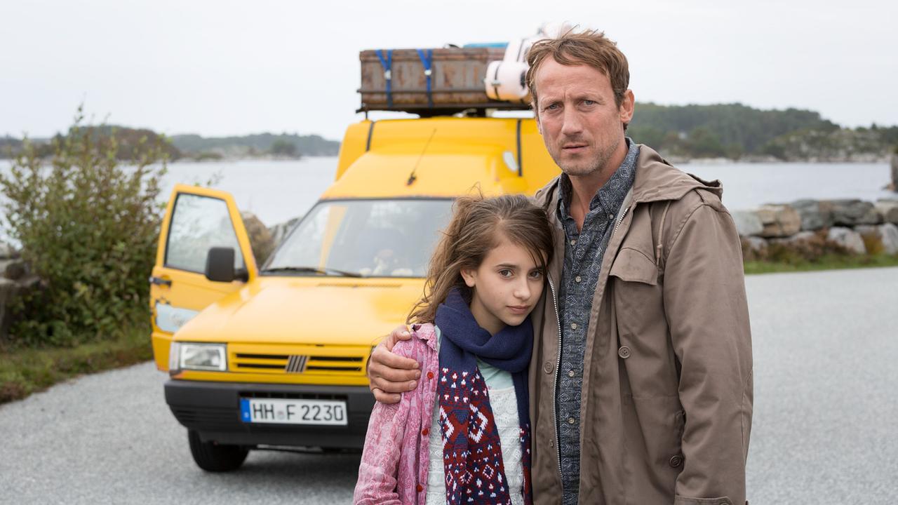 Ein Mann und ein Mädchen stehen umarmt vor einem gelben Auto mit Gepäck auf dem Dach, im Hintergrund Wasser und Küstenlandschaft
