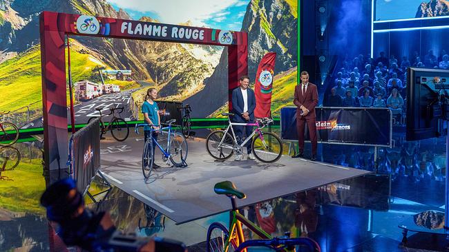 Die 13-jährige Nele Marie im Tour-de-France-Duell mit Jan Ullrich mit ihren Rennräder und Kai Pflaume stehen unter einem Banner „Flamme Rouge“ vor einer Berglandschaftskulisse
