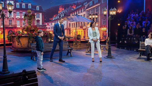 © ORF/NDR/Thorsten Jander Max (10), Kai Pflaume und Jana Pareigis beim Rathaus-Duell in einer Studioszene, die wie ein nächtlicher Stadtplatz gestaltet ist, mit Sitzbänken und Laternen.