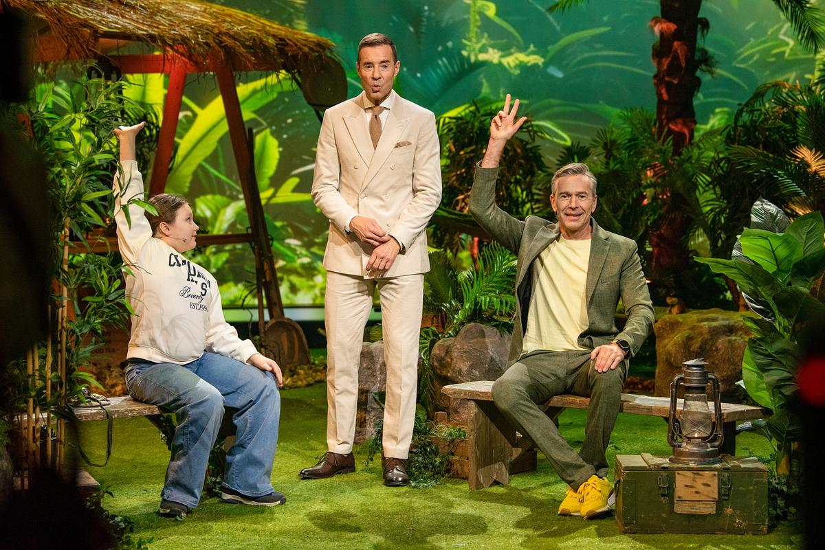 Ella und Dirk Steffens sitzen auf einer Bühne mit Dschungel-Thema und heben die Hand, während Kai Pflaume in der Mitte steht.