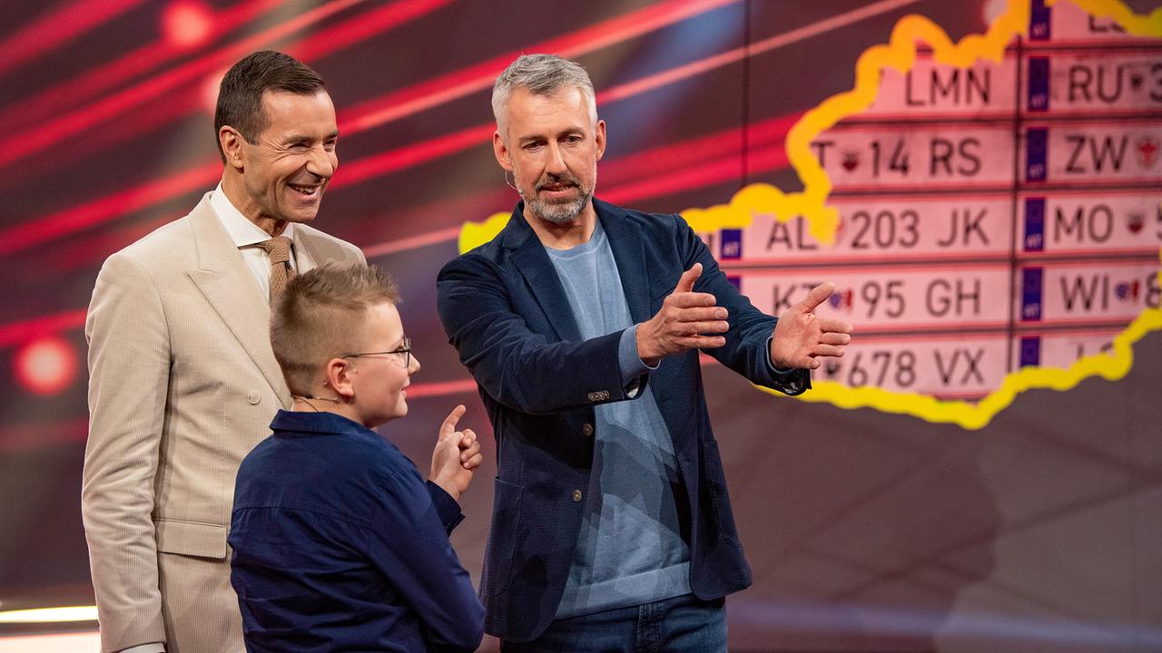 Kai Pflaume, Kurt und Sebastian Pufpaff stehen beim Auto-Kennzeichen-Duell vor einer Tafel mit Nummernschildern und unterhalten sich gestikulierend.