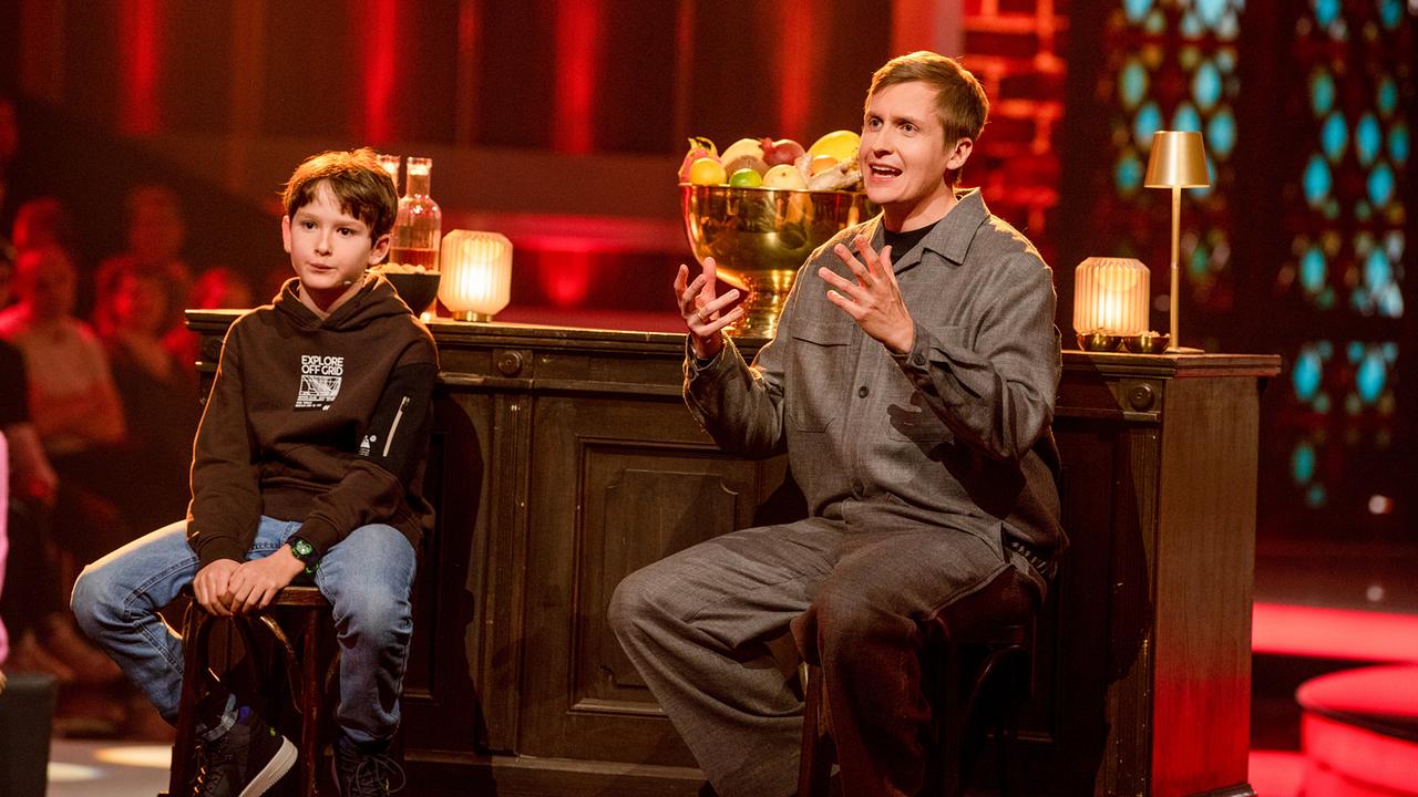 Theodor aus Graz und Till Reiners sitzen beim Sketche-Duell an einer Bar auf einer Bühne, umgeben von warmem rotem Licht.