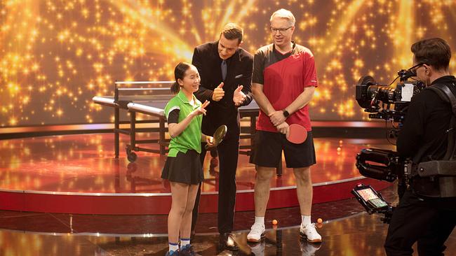 © ORF/NDR/Thorsten Jander Beim Tischtennis‑Duell stehen Enya und Frank im Sport-Outfit mit Schlägern auf der hell beleuchteten Bühne, zwischen ihnen steht Kai Pflaume und vor ihnen ein Kameramann.