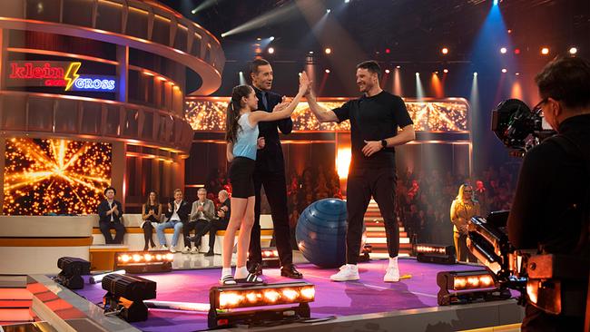 © ORF/NDR/Thorsten Jander Beim Ball-Seilsprung-Duell geben sich Emma und Dario Cologna auf der Bühne ein High‑Five. Moderator Kai Pflaume steht neben ihnen, während das Publikum und ein Kamerateam zuschauen.