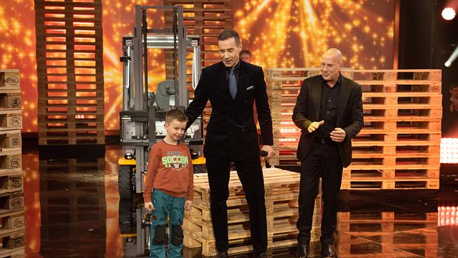 © ORF/NDR/Thorsten Jander Auf einer Showbühne mit Paletten und Gabelstapler stehen Tim, Kai Pflaume und Heino Ferch inmitten einer industriell inszenierten Kulisse.