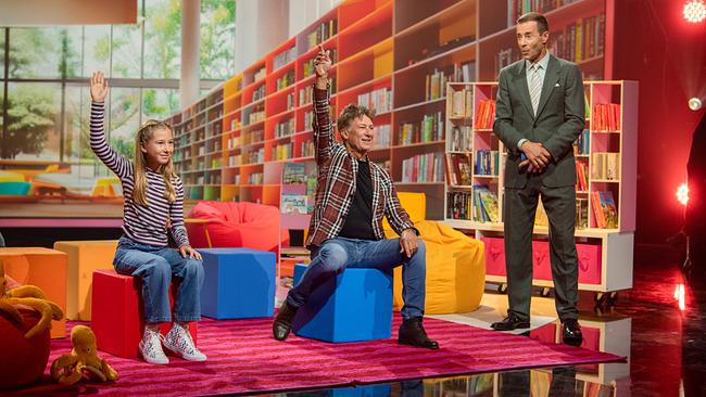 © ORF/NDR/MBTV - Produktions GmbH/Thorsten Jander Marlies (12) und Tobias Moretti sitzen beim Kinderbuch-Duell in einer bunt gestalteten Bibliothekskulisse und zeigen auf, während Kai Pflaume daneben moderiert.