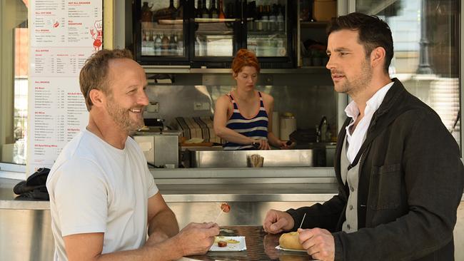 Chefinspektor Max Steiner im weißen T-Shirt und Inspektor Felix Burger in weißem Hemd, grauem Gilet und dunkler Jacke am Würstelstand von Swobo im blau-weiß gestreifem Träger-Shirt