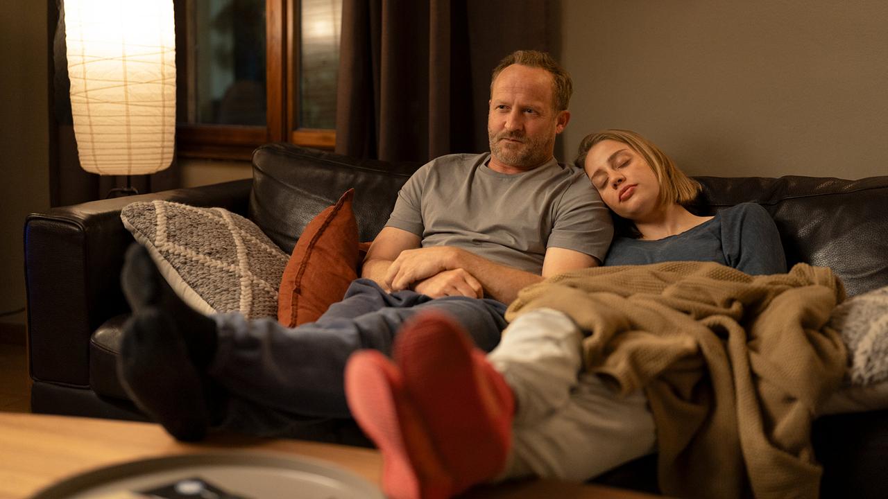 Kommissar Max Steiner sitzt mit Anna Lang, die sich an seine Schulter lehnt, abends auf einem Sofa in einem gemütlich beleuchteten Wohnzimmer, eingekuschelt in Decken und Kissen.