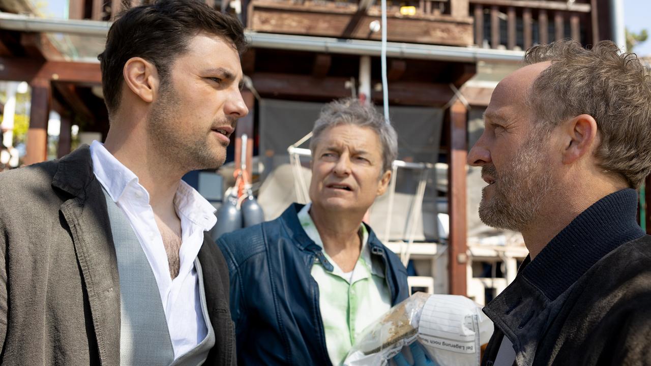 Felix und Max unterhalten sich vor einem Haus mit Holzbalkon, hinter ihnen steht Pathologe Dr. Wippler und hält ein Beweisstück im Plastiksack in der Hand