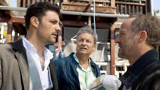 Felix und Max unterhalten sich vor einem Haus mit Holzbalkon, hinter ihnen steht Pathologe Dr. Wippler und hält ein Beweisstück im Plastiksack in der Hand