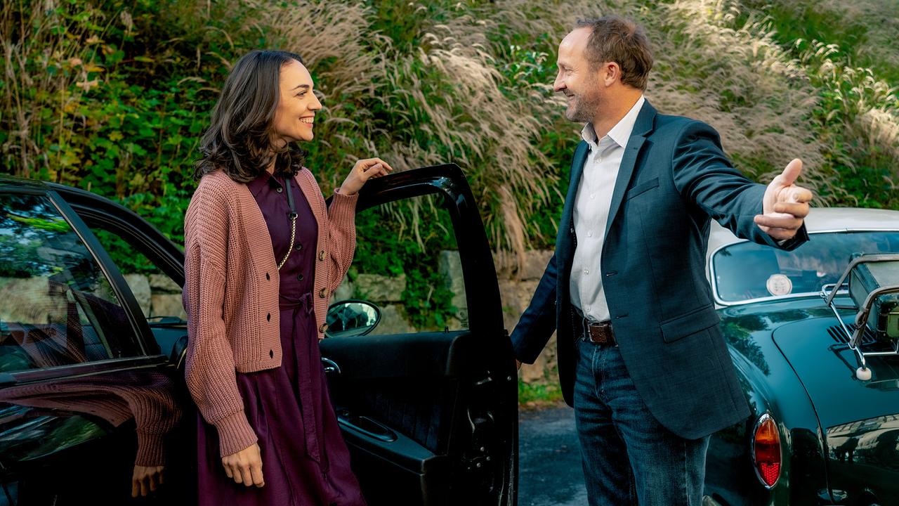 Max Steiner öffnet für Julia die Beifahrertür seines Wagens, beide sind für einen sommerlichen Theaterbesuch bekleidet und lächeln sich gegenseitig freundlich an