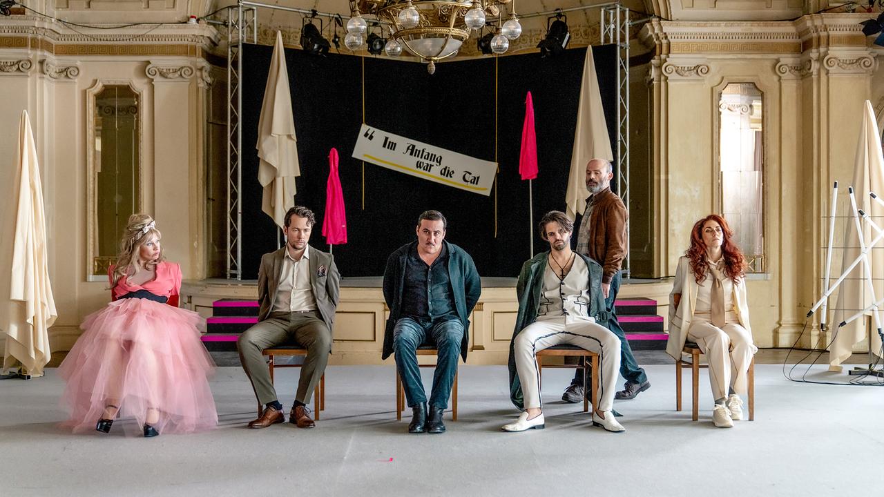 Zwei Schauspielerinnen und drei Schauspieler sitzen an Stühlen gefesselt auf der Bühne eines Theaters, hinter ihnen geht ein bärtiger Mann vorbei, am Bühnenbild ein Schild mit der Aufschrift: Im Anfang war die Tat