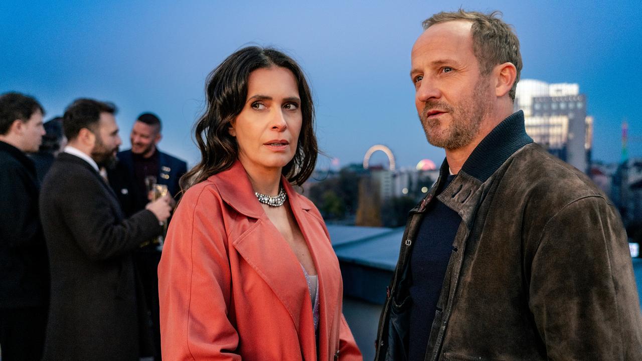 Majaron Evelyn Leitner und Chefinspektor Max Steiner unterhalten sich bei einer Abendveranstaltung auf einer Terrasse miteinander, beide blicken zur Seite in dieselbe Richtung, entfernt im Hintergrund ist das Wiener Riesenrad zu sehen 