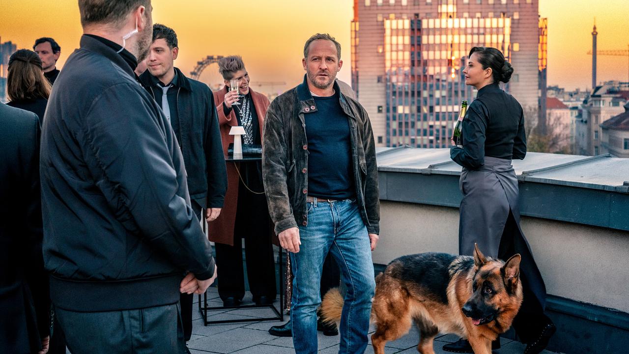 Auf einer Terrasse bei einer Abendveranstaltung mischt sich Chefinspektor Max Steiner mit Polizeihund Rex unter die Gäste, mit anderen Security-Manager sorgt er für Sicherheit, entfernt im Hintergrund ist das Wiener Riesenrad zu sehen