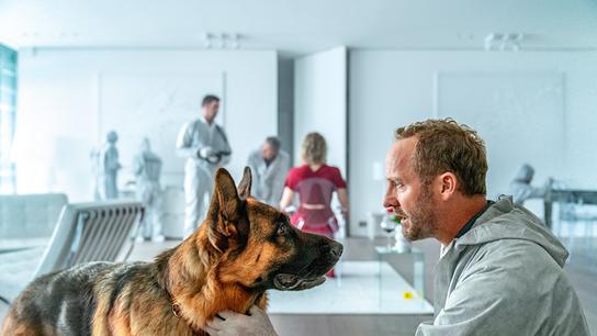 Chefinspektor Max Steiner in einem Schutzanzug der Spurensuche beugt sich zu Schäferhund Rex und schaut ihm in die Augen, im Hintergrund im weißen Appartment eine Frau in rotem Kleid sitzend, Personen in Schutzbekleidung sichern die Spuren am Tatort