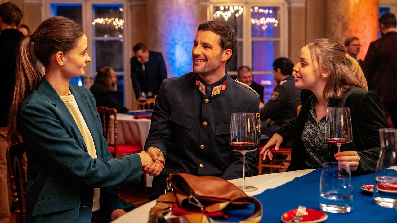 Inspektor Felix in festlicher Uniform bei einer Abendveranstaltung, am Stehtisch von Anna begrüßt er deren Freundin Laura 
