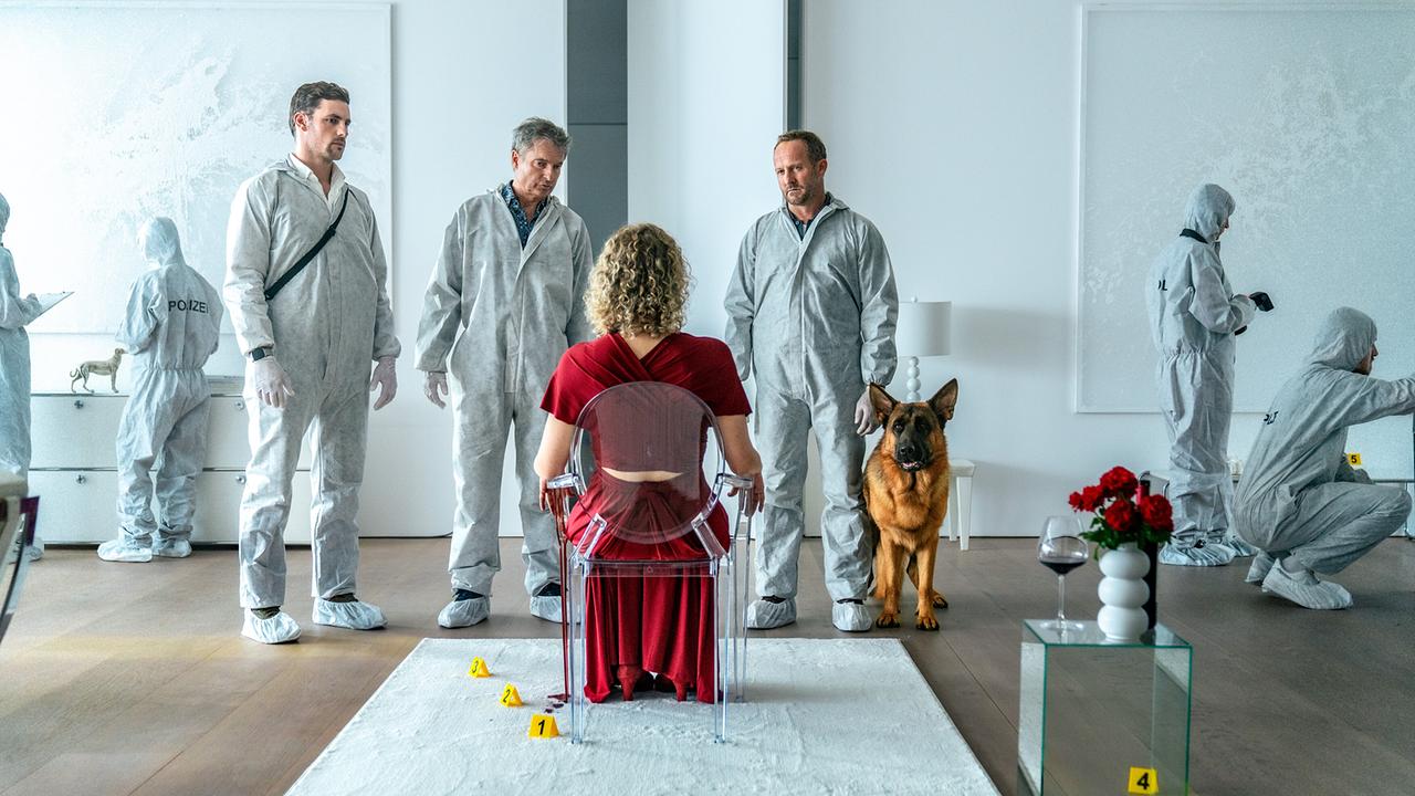In einem hellen weißen Raum sitzt das Mordopfer im roten Kleid auf einem Stuhl, neben ihr ein Glastisch mit Rosen und einem Glas Rotwein, Felix, Max und Dr. Wippler in Schutzbekleidung unterhalten sich miteinander am Tatort, Schäferhund Rex steht neben ihnen