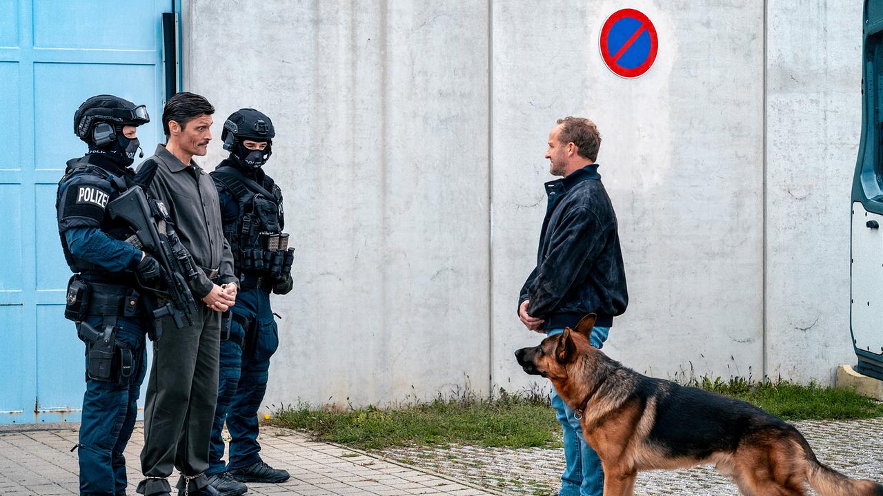 Der Häftling Harald Binder wird in Handschellen von zwei Polizisten mit Helmen und Schutzbekleidung begleitet, vor einer Betonmauer begegnet ihm Chefinspektor Max mit Schäferhund Rex an seiner Seite