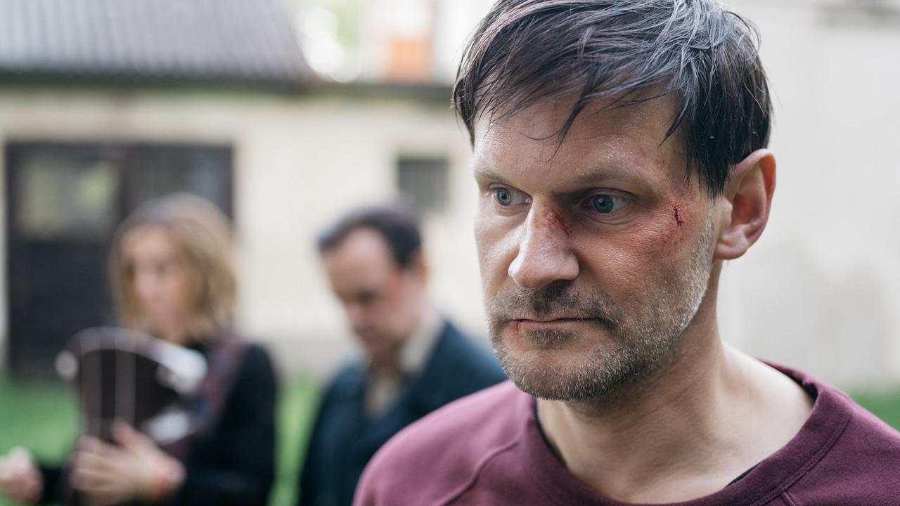 © ORF/Superfilm/Ingo Pertramer Nahaufnahme von Robert Schmatz in einem burgunderroten Sweatshirt mit Schürfwunden im Gesicht auf einer Wiese vor Häusern
