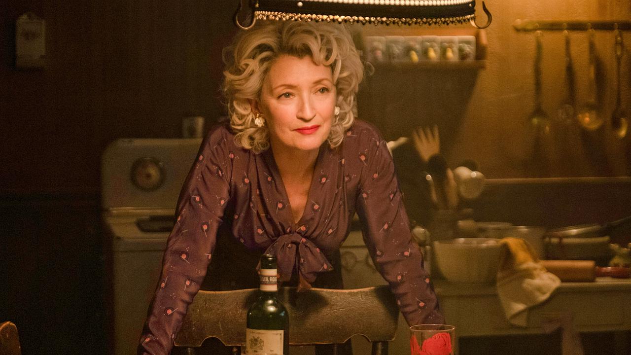 © ORF/Universal/Focus Features/Kimberly French In einer dunklen Küche lehnt sich eine elegant gekleidete, blondhaarige Frau über die Sessellehne auf den Tisch und blickt ihr Gegenüber freundlich an, auf dem Tisch eine Weinflasche