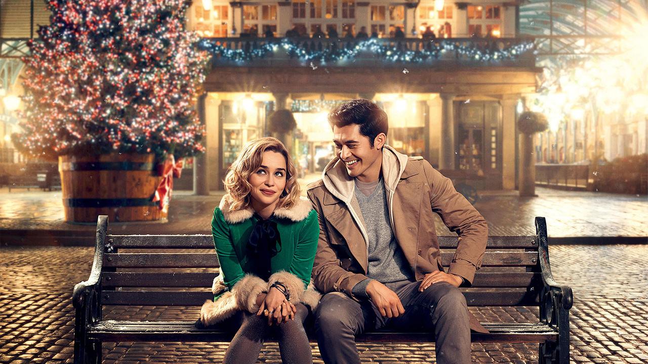 Emilia Clarke sitzt als Kate in grüner Winterjacke mit Fellbesatz mit Henry Golding als Tom in brauner Jacke auf einer Bank vor festlich beleuchtetem Weihnachtsbaum und geschmücktem Gebäude