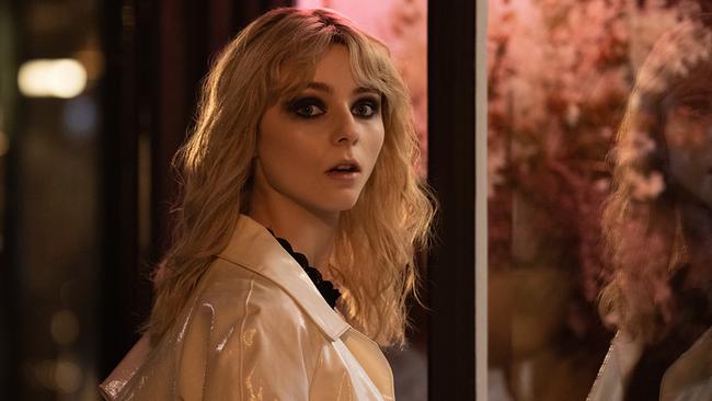 Eine blondhaarige, junge Frau in einem weißen Mantel, steht in der Nacht auf vor einer beleuchteten Auslageund blickt erstaunt über ihre Schulter zurück