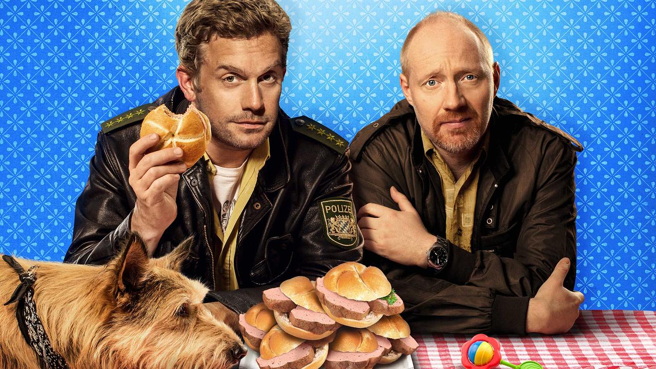 Ein Mann in Polizeijacke und ein Mann in brauner Jacke sitzen vor einem Stapel Leberkässemmeln, einer isst gerade, vor ihnen liegt ein Hund am Tisch mit Blick auf das Essen