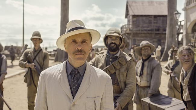 Christoph Waltz als Capt. Léon Rom in weißem Anzug mit blauem Hemd und blauer Krawatte und weißem Hut mit schwarzer Krempe, hinter ihm bewaffnete Männer - alle blicken in dieselbe Richtung nach vorne