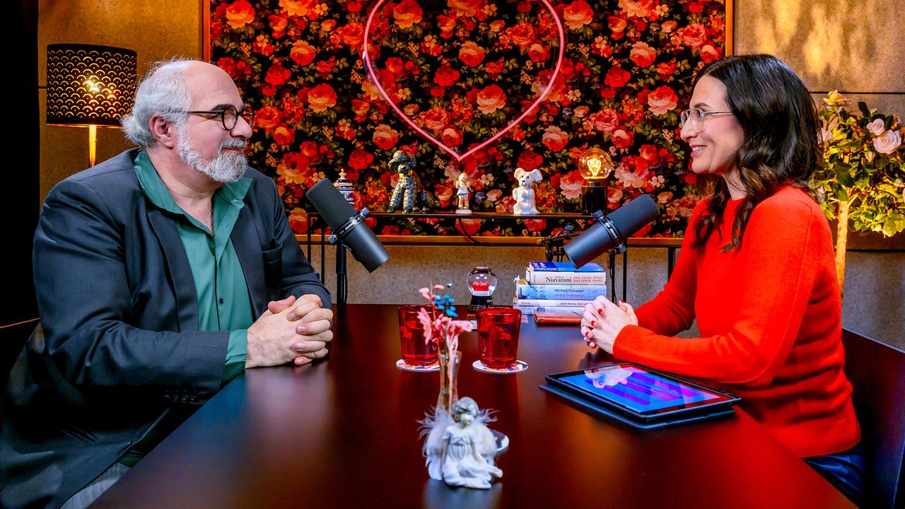 Michael Niavarani und Nina Horowitz sitzen beim Gespräch an einem dunklen Holztisch beim ORF-„Liebesg’schichten und Heiratssachen – Der Podcast“