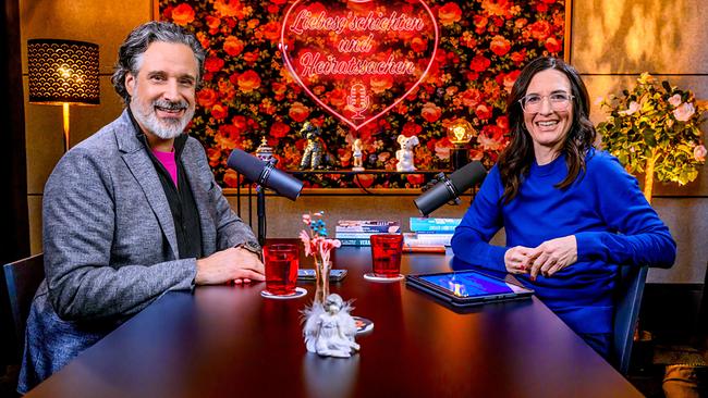 Pablo Hagemeyer und Nina Horowitz sitzen sich in einem Podcast-Studio mit Mikrofonen gegenüber an einem Tisch vor einer floralen Wand mit Herz-Neonschild mit der Aufschrift „Liebesgeschichten und Heiratssachen“ und lächeln in die Kamera.