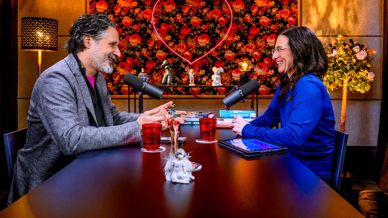 Pablo Hagemeyer und Nina Horowitz sitzen sich in einem Podcast-Studio mit Mikrofonen gegenüber und sprechen an einem Tisch vor einer floralen Wand mit Herz-Neonschild mit der Aufschrift „Liebesgeschichten und Heiratssachen“.