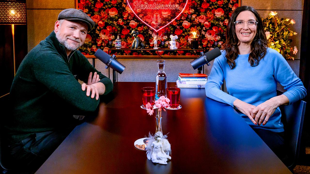 Thomas Stipsits und Nina Horowitz sitzen an einem Tisch mit Mikrofonen in einem gemütlichen Studio, vor einer floralen Wand mit leuchtendem Herzmotiv und der Aufschrift „Liebesgeschichten und Heiratssachen“.