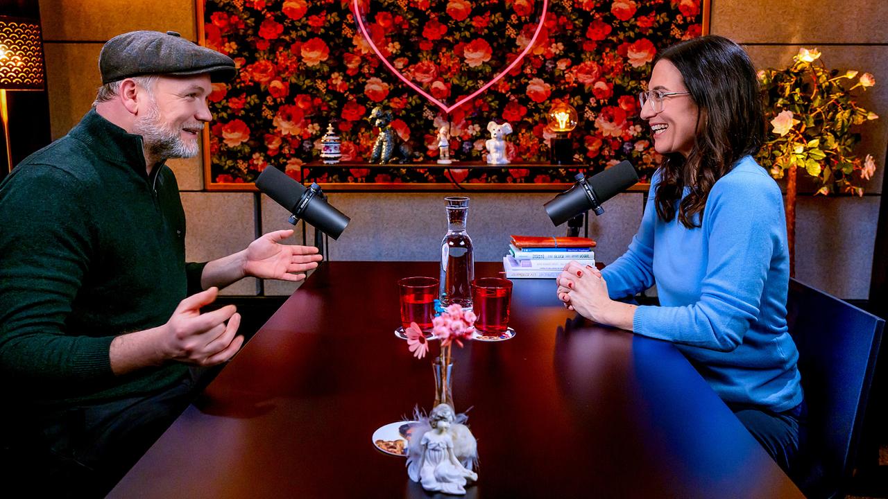 Thomas Stipsits und Nina Horowitz sitzen im angeregten Dialog an einem Tisch mit Mikrofonen in einem gemütlichen Studio, vor einer floralen Wand mit leuchtendem Herzmotiv und der Aufschrift „Liebesgeschichten und Heiratssachen“.