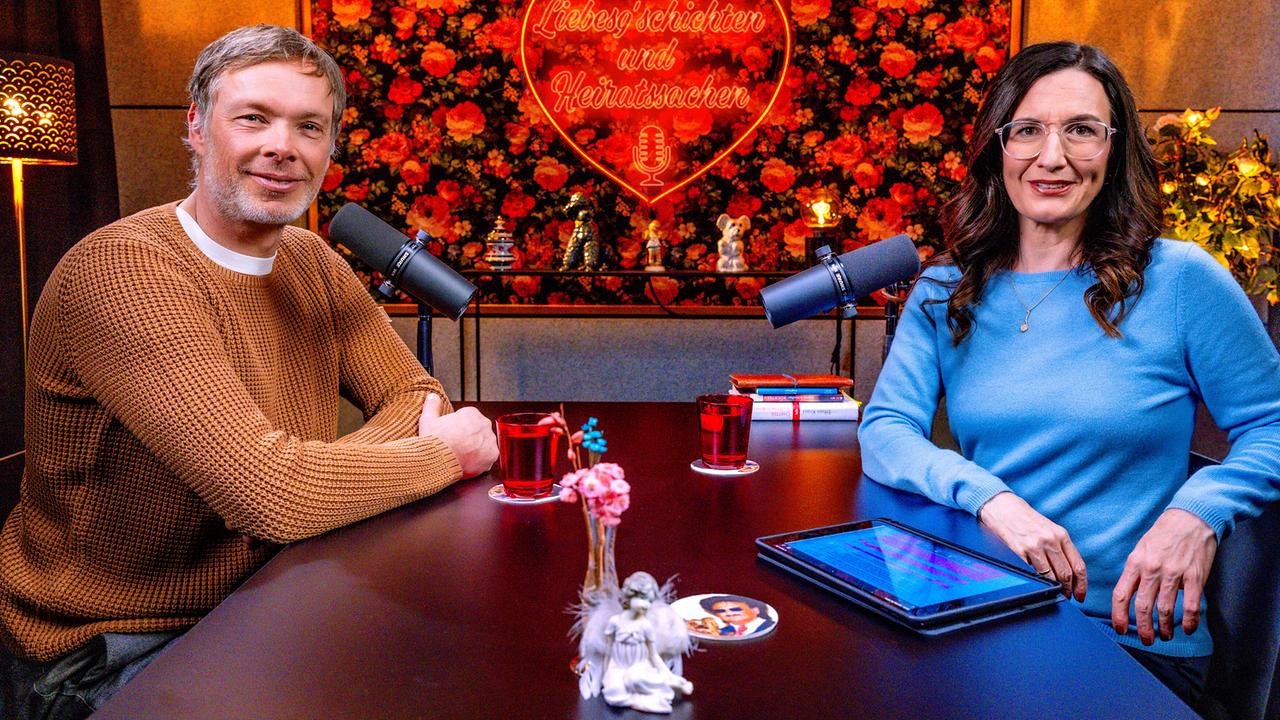 © ORF/Hans Leitner Thomas Sykora und Nina Horowitz sitzen an einem Tisch mit Mikrofonen in einem gemütlichen Studio, vor einer floralen Wand mit leuchtendem Herzmotiv und der Aufschrift „Liebesgeschichten und Heiratssachen“.