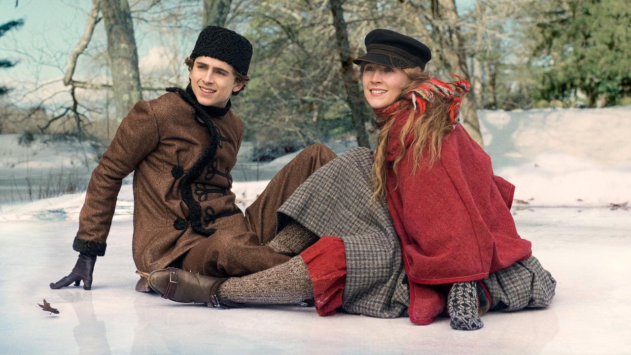 Theodore und Saoirse sitzen sich in historischer Kleidung beim Eislaufen auf dem Eis gegenüber und blicken freundlich begrüßend in dieselbe Richtung