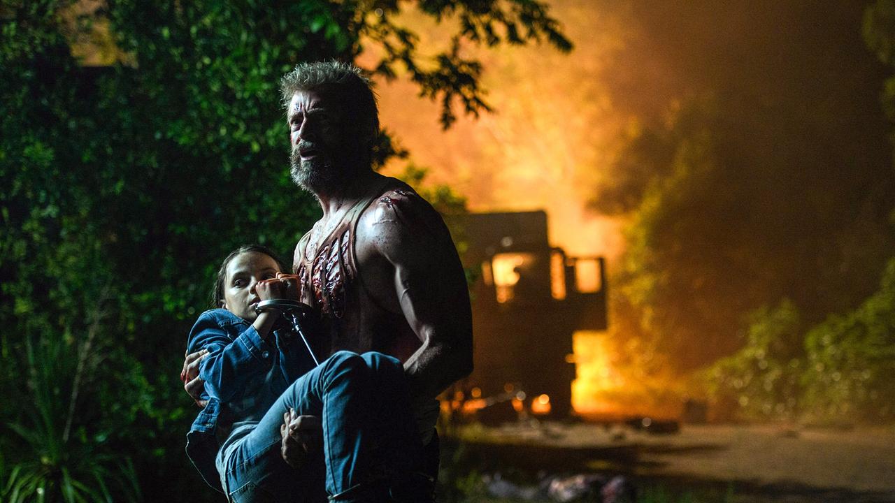 Hugh Jackman als Wolverine trägt Dafne Keen als das Mädchen Laura Kinney, auf der Sraße, die durch ein Waldstück führt, brennt im Hintergrund ein Lieferwagen