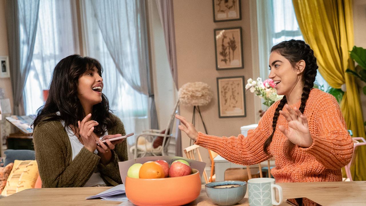 Zwei junge Frauen sitzen in einem hellen Wohnzimmer an einem Tisch, während eine auf ihrem Handy tippt schauen sich die beiden einander an und lachen