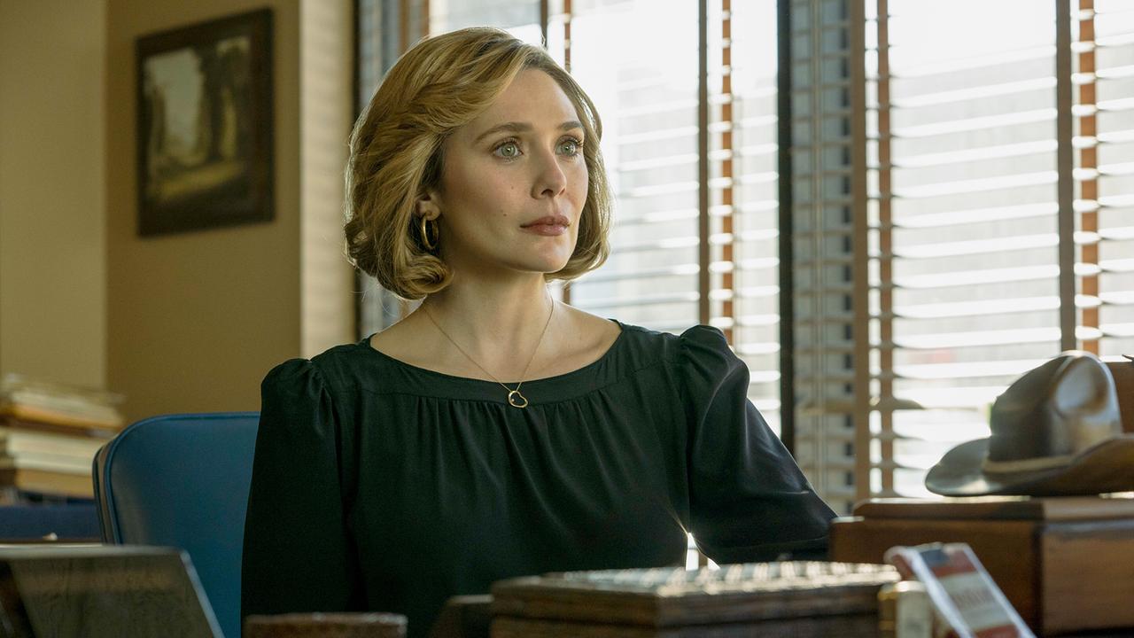 Elizabeth Olsen als Candy Montgomery, in einem dunkelgrünen Kleid sitzt an einem Schreibtisch und blickt ihrem Gegenüber ernst und gefasst entgegen