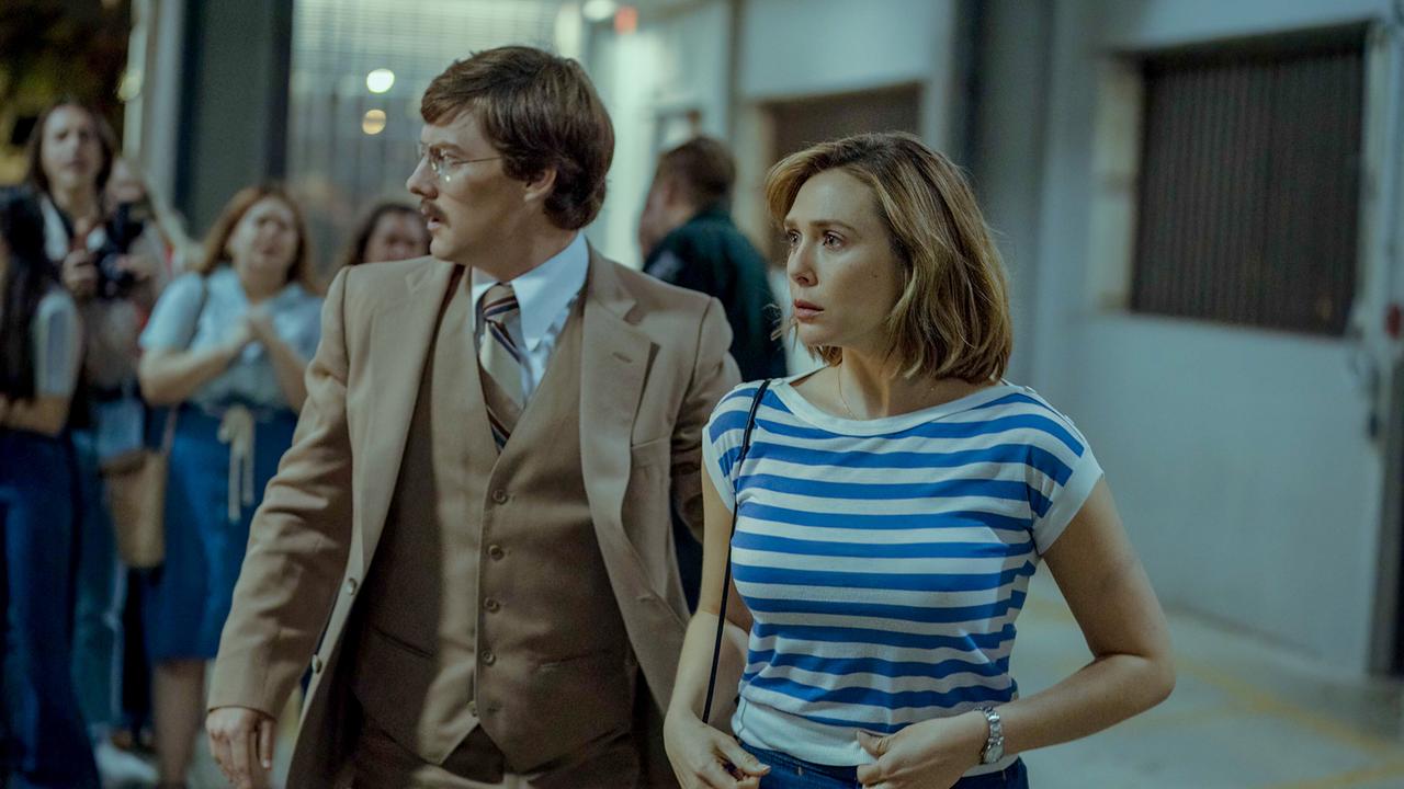 © ORF/Warner/HBO Max Eine Frau in blau-weiß gestreiftem Shirt und ein Mann in beigefarbenem Anzug gehen durch einen hell beleuchteten Raum, im Hintergrund stehen Menschen und schauen betroffen die beiden an