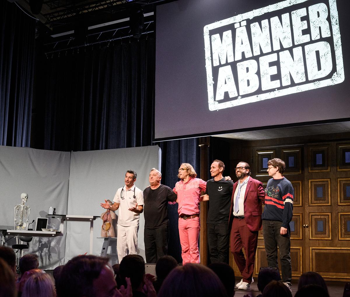 Omar Sarsam, Andreas Vitásek, Blonder Engel, Gernot Kulis, Gerald Fleischhacker und Christoph Fritz stehen bei der Aufführung von „Männerabend“ gemeinsam auf der Theaterbühne vor dem Publikum.