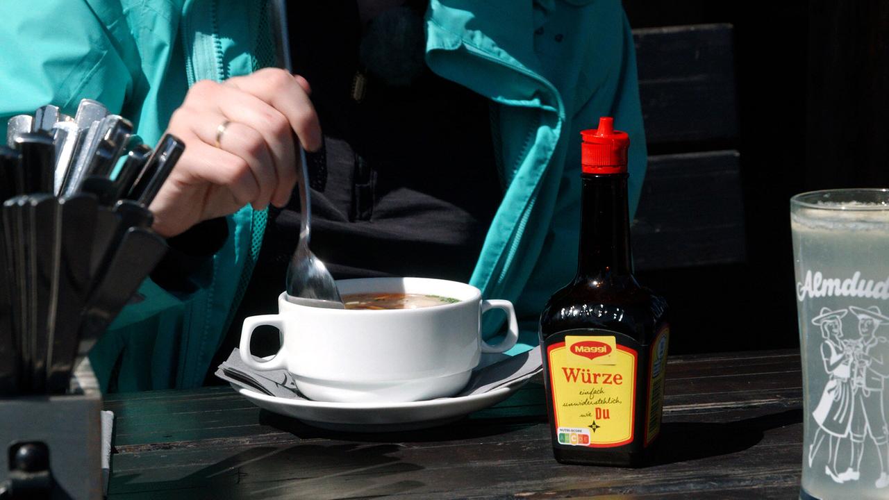 © ORF/Bernhard Werany Eine Person in türkiser Jacke und schwarzem Shirt sitzt an einem Tisch, sie taucht einen Löffel in eine Suppentasse neben der auf dem Tisch eine Flasche Speisewürze steht