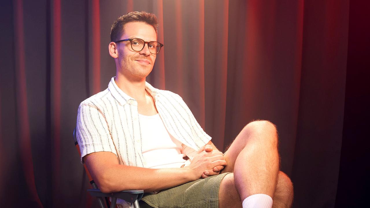 Martin Frank sitzt auf der Bühne in einem Theater mit Vorhängen im Hintergrund, sommerlich gekleidet mit kurzärmeligem Hemd, Shorts, Socken und Badeschlappen.