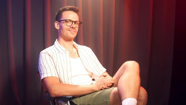 Martin Frank sitzt auf der Bühne in einem Theater mit Vorhängen im Hintergrund, sommerlich gekleidet mit kurzärmeligem Hemd, Shorts, Socken und Badeschlappen.