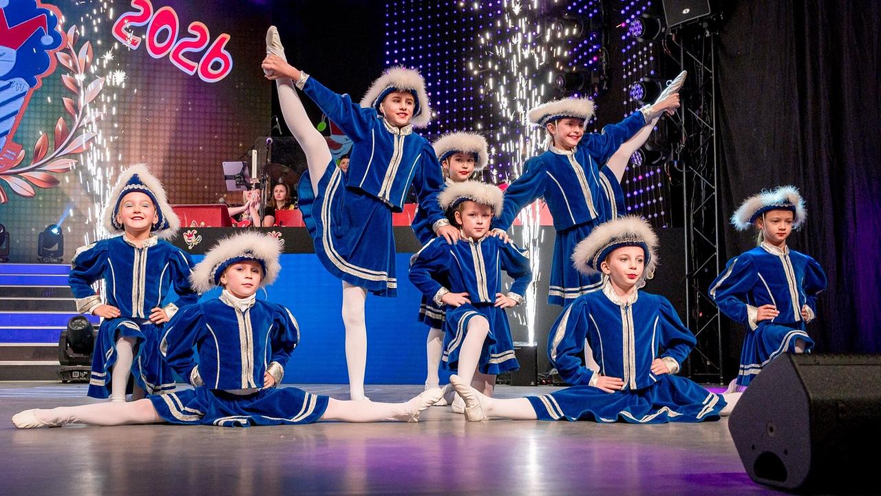 © ORF/Villacher Fasching/Knaus Die kleine Garde in blauen, pelzbesetzten Kostümen zeigt eine akrobatische Tanzpose auf der "Mini Lei Lei"-Bühne, während im Hintergrund Funkenfontänen und die Jahreszahl „2026“ zu sehen sind.
