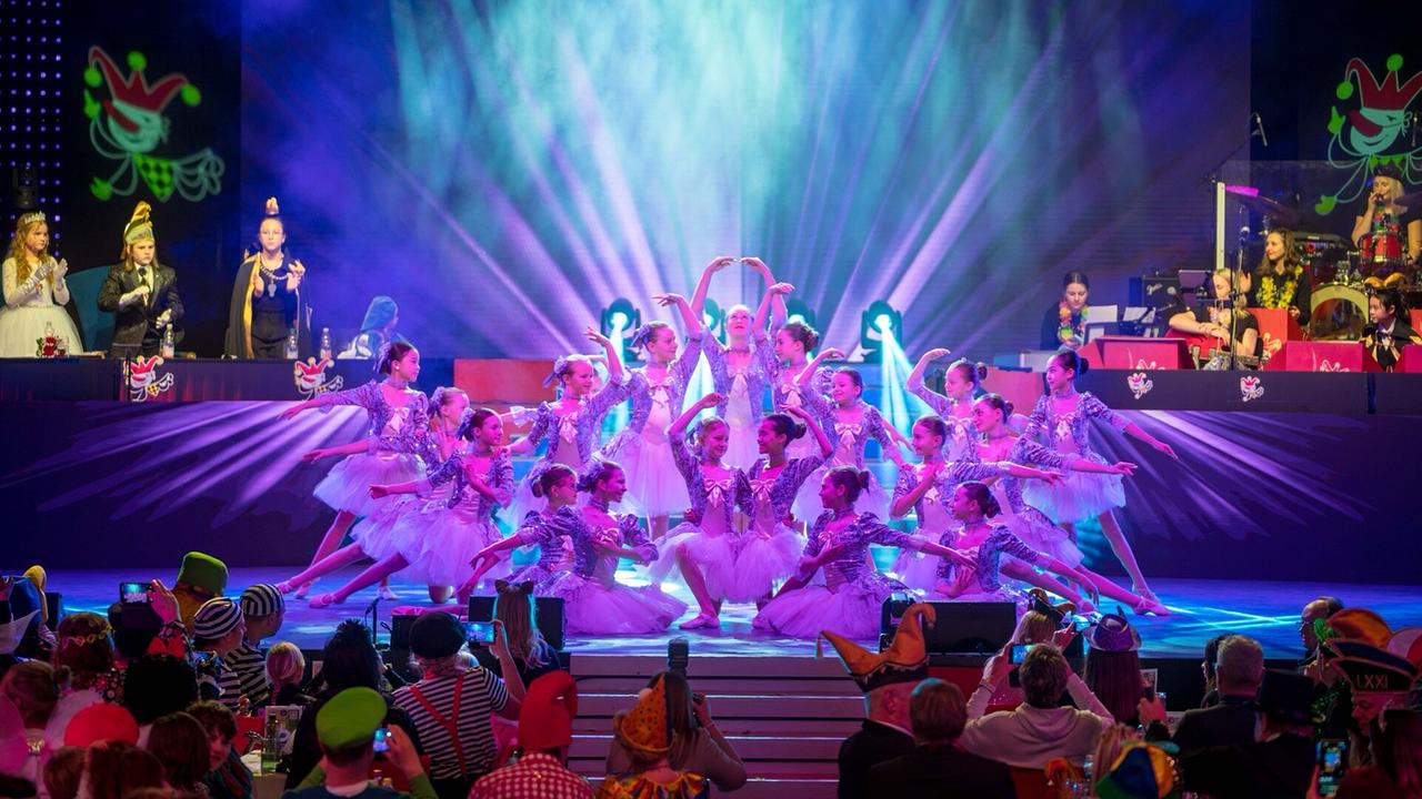 © ORF/Villacher Fasching Auf einer farbig ausgeleuchteten Bühne führen zahlreiche Kinder in glitzernden Kostümen eine Ballett-Choreografie vor einem kostümierten Publikum auf.