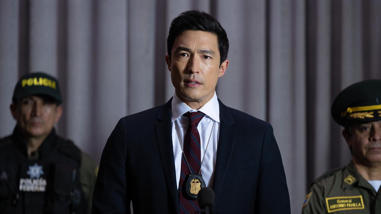 © ORF/Sony Pictures Entertainment/Temma Hankin Daniel Henney als Agent Park bei einer Pressekonferenz, er trägt einen dunklen Anzug, weißes Hemd und dunkelrote Krawatte, an einer Kette hängt seine Polizeimarke, neben ihm stehen zwei uniformierte Männer
