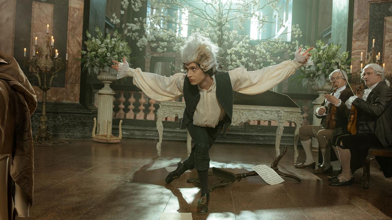 © ORF/WDR/Story House Pictures/Armands Virbulis Wolfgang Amadeus verbeugt sich theatralisch in einem prunkvollen Saal, im Hintergrund sitzen Musiker mit Streichinstrumenten