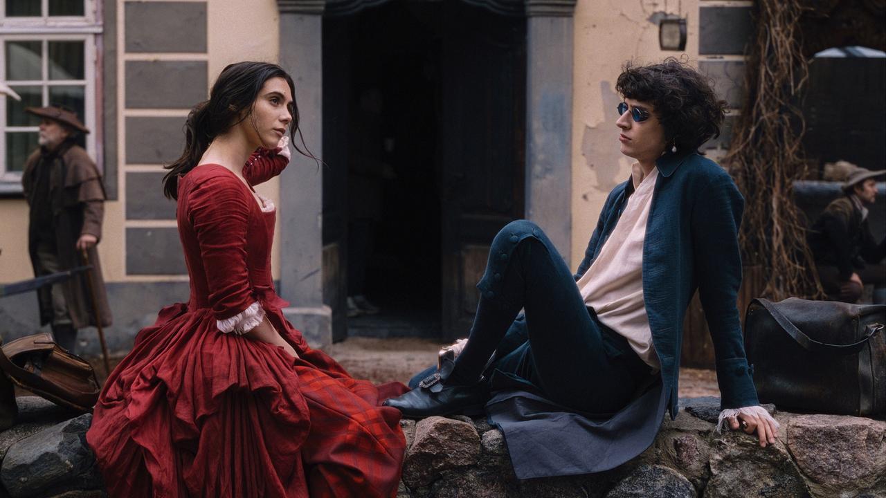 © ORF/WDR/Story House Pictures/Armands Virbulis Maria Anna Mozart in rotem Kleid und Wolfgang Amadeaus Mozart in dunkelblauer Kleidung mit Sonnenbrille sitzen sich auf einer Steinmauer in einer Straße gegenüber