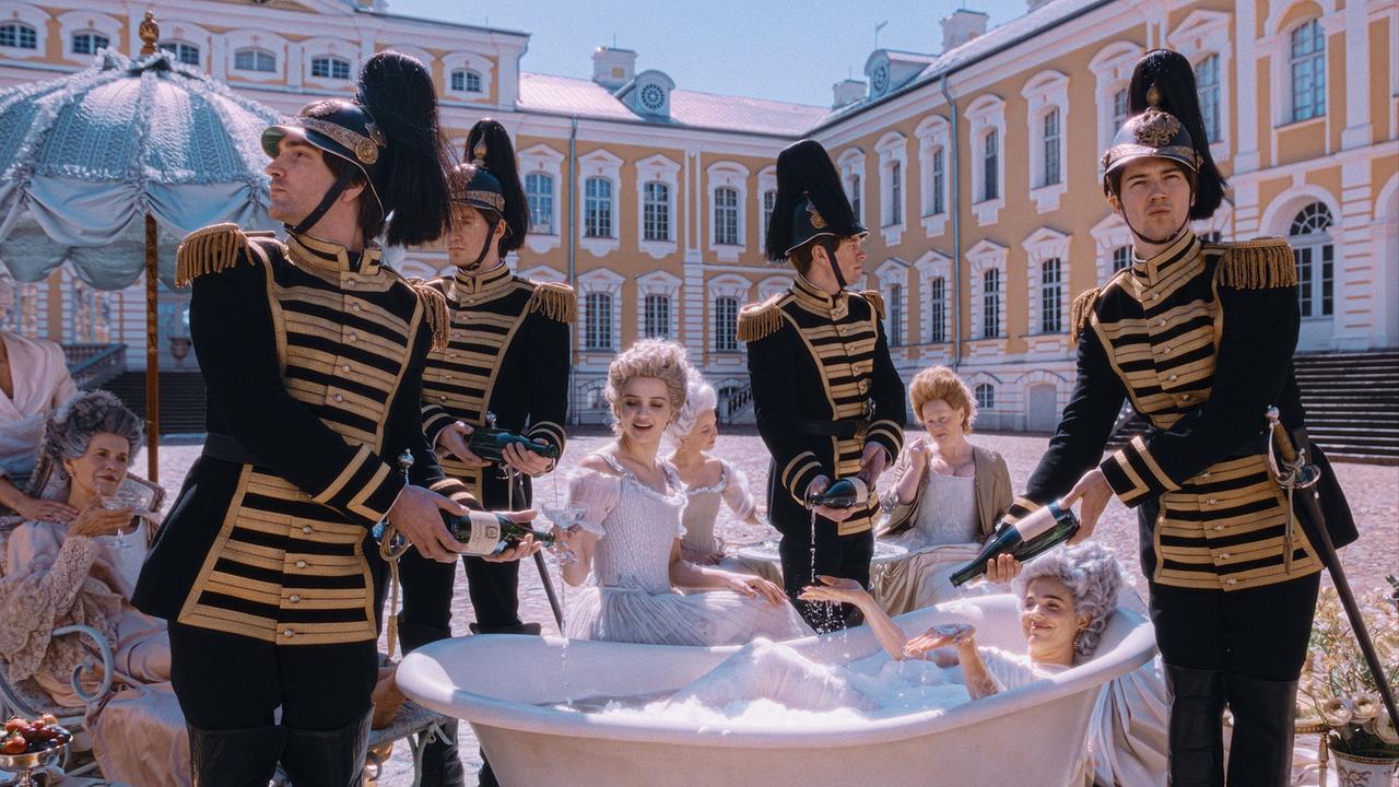 Mehrere Männer in schwarzen Uniformen mit goldenen Verzierungen umringen Verena Altenberger, die als Marie Antoinette in einer Schaum gefüllten Badewanne im Freien vor einem prunkvollen Palast liegt
