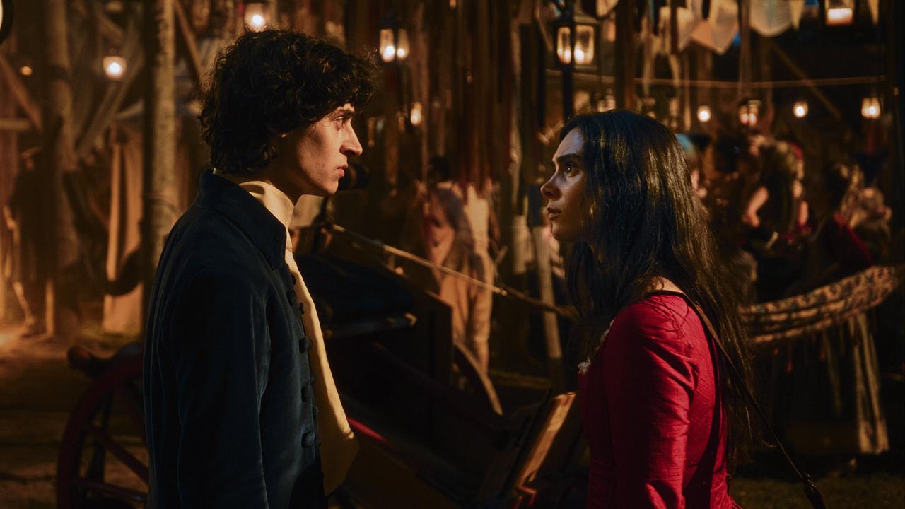 Wolfgang Amadeus in dunkler Kleidung und Anna Maria im roten Kleid stehen sich auf einem nachts belebten Platz gegenüber und blicken sich ernst an